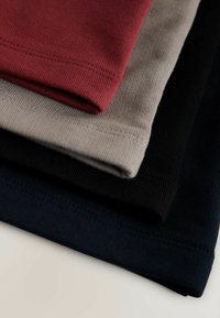 Cuatro camisetas dobladas en rojo, gris, negro y azul marino. La tela tiene una textura acanalada con costuras visibles a lo largo de los bordes.