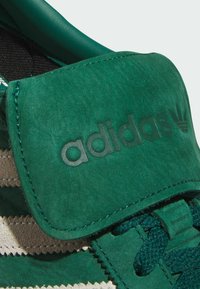 Green suede sneaker featuring a padded tongue with embossed logo, contrasting textile stripes, and lace closure for a secure fit.
