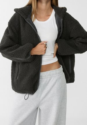 Person iført en mørkegrå tekstureret lynlåstrøje over en hvid cropped tanktop og lysegrå joggingbukser med elastisk talje.