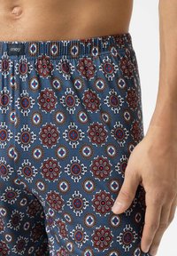 Close-up van een persoon die blauwe boxershorts met een patroon draagt, voorzien van rode, witte en bruine bloemen- en cirkelvormige ontwerpen, met de hand rustend aan de zijkant.
