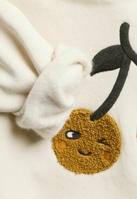 Witte stof met een bruine geborduurde kers met een lachend gezicht, zwarte bladdetail en zachte, textuur elementen.