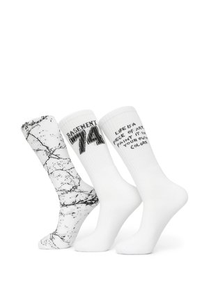 Trzy białe skarpetki typu crew; jedna z czarnym marmurkowym wzorem, jedna z napisem "BASEMENT 74" oraz jedna z częściowym czarnym tekstem motywacyjnym.