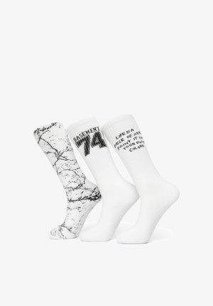 Trzy białe skarpetki typu crew; jedna z czarnym marmurkowym wzorem, jedna z napisem "BASEMENT 74" oraz jedna z częściowym czarnym tekstem motywacyjnym.