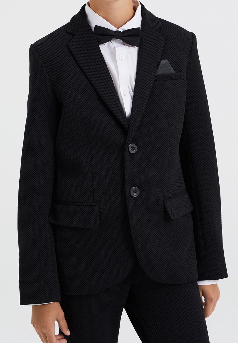 Svart texturerad blazer med två knappar, slagkrage och sidofickor. Matchad med en vit skjorta och en svart fluga. Accenterad med en fickruta.