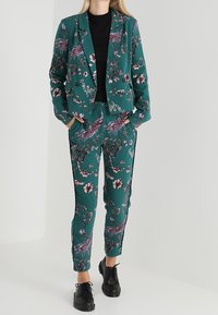 Costume floral vert avec manches longues, blazer ajusté et pantalon fuselé. Comprend un haut noir en dessous et des chaussures noires pour le contraste.