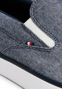 Primer plano de un zapato azul con textura, tipo slip-on, con panel lateral elástico blanco y pequeña etiqueta de tela roja, blanca y azul encima de la suela de goma blanca.