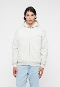 Sweat à capuche zippé gris avec une capuche doublée en peluche blanche, poches avant, détail du logo sur la poitrine gauche, et bords côtelés au niveau des poignets et de l'ourlet.