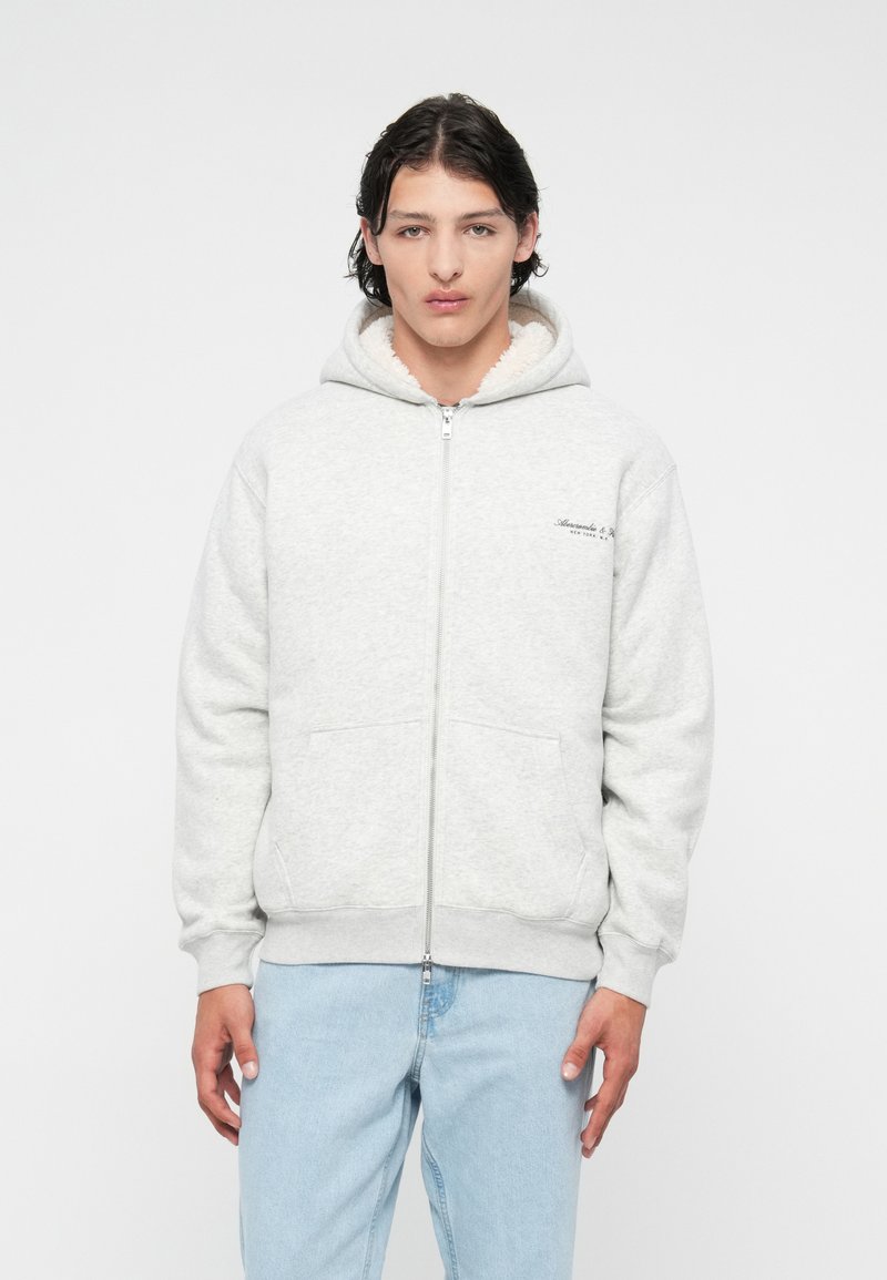 Sweat à capuche zippé gris avec une capuche doublée en peluche blanche, poches avant, détail du logo sur la poitrine gauche, et bords côtelés au niveau des poignets et de l'ourlet.