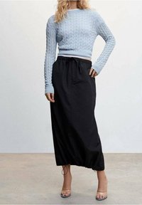 Femme portant un pull court en maille torsadée bleu clair, une jupe midi noire à cordon de serrage et des sandales à talons transparents, debout sur un sol en béton.