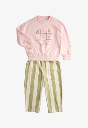 Felpa rosa chiaro con design grafico oro e rosa, abbinata a pantaloni a righe crema e verdi e sneakers marroni. Berretto in maglia rosso sulla testa.