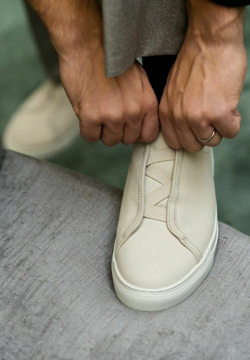 Beige leather sneaker with elastic laces, flat sole, and textured detailing. Hands are tying the laces, placed on a soft, gray surface.