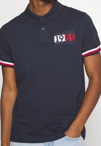 Navyblauwe polo met kraag, voorzien van rood, wit en blauwe gestreepte mouwaccenten en geborduurde "HILFIGER 1985 NEW YORK" branding.