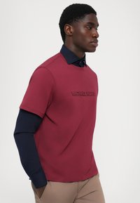 Burgundy kortærmet skjorte med prægede "Michael Kors" logo, stylet over en navy langærmet skjorte, parret med tan bukser.