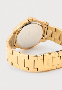 Zlata ura iz nerjavečega jekla z gladkim zaključkom, povezano zapestnico in okroglo ohišje. Na zadnji strani so prikazane blagovna znamka in podatki o vodoodpornosti.