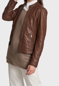 Hnědá kožená bomber bunda se zaobleným límcem, zipovými kapsami a přiléhavými rukávy, nošená přes béžový svetr a bílé kalhoty.