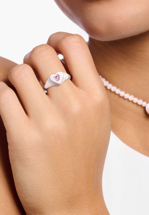 White heart-shaped ring with a pink gemstone center. Smooth, glossy finish with subtle wavy details. Accompanied by a pink bead necklace.