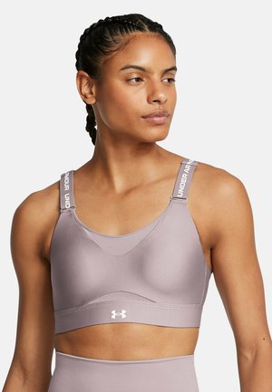 Mujer que lleva un sujetador deportivo malva con tirantes ajustables de "Under Armour", de pie contra un fondo blanco liso, mirando hacia un lado.