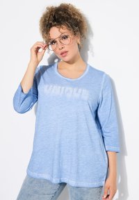 Světle modré tričko s dlouhým rukávem, na kterém je v texturovaném bílém písmu napsáno "UNIQUE". Má volný střih a měkkou tkaninu.