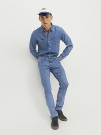 Camisa de mezclilla y jeans en azul claro, con dos bolsillos en el pecho y cierre de botones. Zapatillas negras y gorra crema con texto azul.