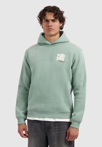 Man met krullend haar, gekleed in een lichtgroene hoodie met opdruk op de borst, gedragen over een wit shirt en donkere jeans, staand in een neutrale houding.