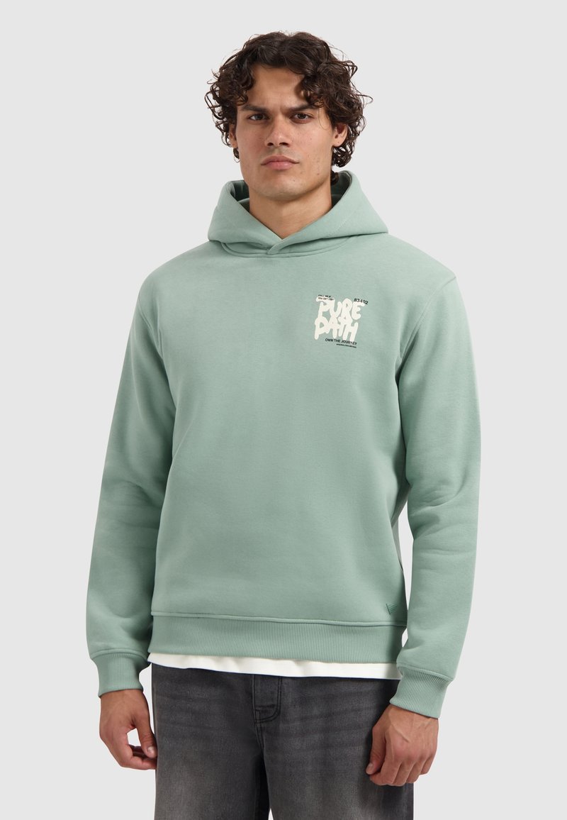 Man met krullend haar, gekleed in een lichtgroene hoodie met opdruk op de borst, gedragen over een wit shirt en donkere jeans, staand in een neutrale houding.