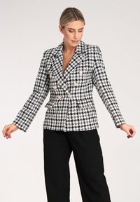 Czarny i biały żakiet w kratę o podwójnym zapięciu, złotych guzikach, wyraźnych ramionach i długich rękawach. Materiał wydaje się teksturowany.