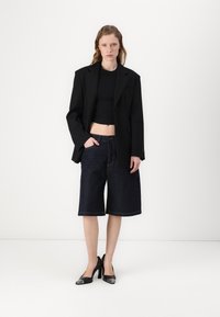 Črn oversized blazer preko skrajšanega črnega topa, v kombinaciji s temnimi denim kratkimi hlačami in črnimi visokimi petami s kovinskimi detajli.