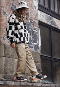 Černobíle kostkovaná fleecová bunda, khaki kalhoty a černé tenisky s oranžovými detaily. Městské pozadí s cihlovou zdí.