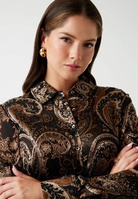 Giovane donna con lunghi capelli castani che indossa orecchini a cerchio dorati e una camicia a bottoni con motivo paisley in nero, marrone e beige, con le braccia incrociate.