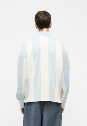 Férfi világoskék-fehér, függőleges csíkos hosszú ujjú galléros ingben, laza sötét farmernadrágban, háttal áll.