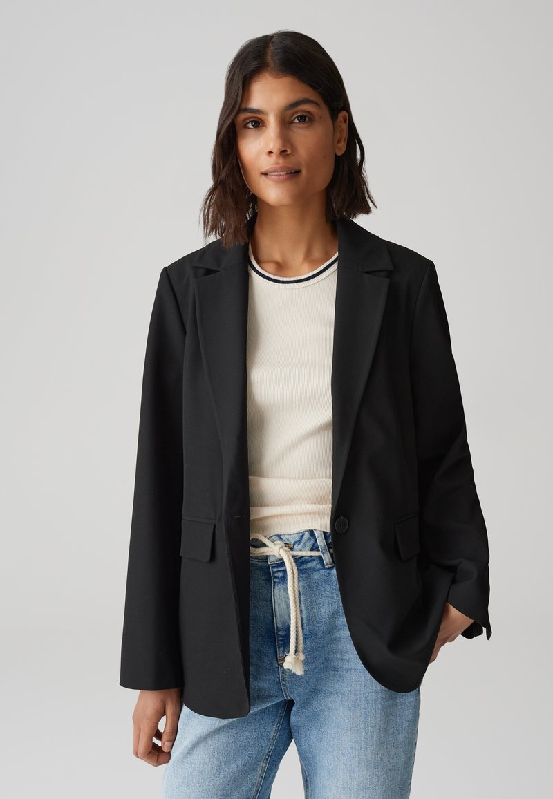 Zwarte blazer met een getailleerde pasvorm, notch revers, enkele knoopsluiting en voorzakken, gedragen over een licht geribbeld topje en blauwe jeans.