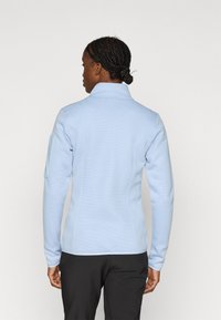 Ανοιχτό μπλε φλις μπουφάν με ψηλό γιακά, υφή υφάσματος και εφαρμοστή σχεδίαση. Διαθέτει πλαϊνή τσέπη στο αριστερό μανίκι και ελαστικά μανσέτες.