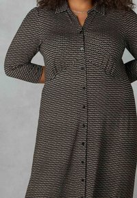 Musta ja kerma värinen kuvioitu mekko, jossa on kaulus, nappikiinnitys ja kolme neljännestä hihaa; siinä on hienovarainen geometrinen muotoilu.