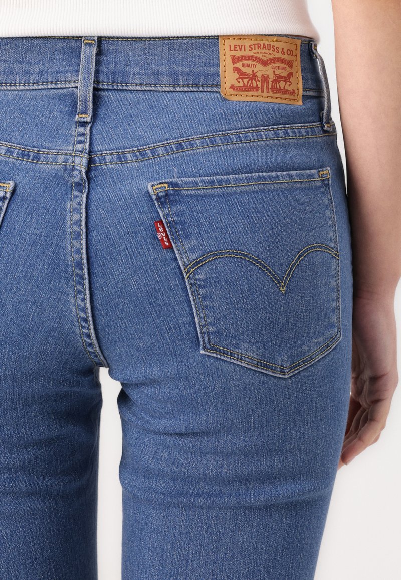 Jean en denim bleu au design ajusté, avec une patch en cuir au niveau de la ceinture et des poches arrières courbes distinctives avec des surpiqûres jaunes.
