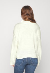 Kobieta z długimi brązowymi włosami, ubrana w biały sweter i niebieskie dżinsy, stojąca tyłem na jednolitym jasnym tle.