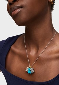 Srebrny naszyjnik z łańcuszkiem ozdobiony różnorodnymi zawieszkami: turkusową koralikiem, teksturowanymi srebrnymi wzorami oraz złotym akcentem. Noszony na ciemnoniebieskiej bluzce.