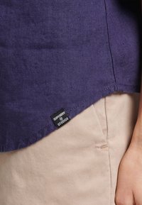 Närbild av en person som bär en lila linneskjorta med en "Superdry Studios"-etikett och beiga byxor, handen hvilar bredvid fickan på byxorna.