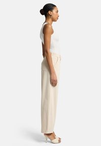 Beige Hochbundhosen mit lockerer Passform, ausgestattet mit Seitentaschen, kombiniert mit einem weißen, figurbetonten Tanktop und beige offenen High Heels.