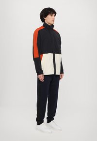 Vindjacka med hög krage, flerfärgad design i svart, orange och gräddfärg. Matcher med marinblå joggers och vita sneakers.