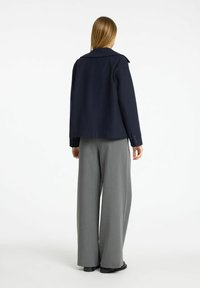Giacca in misto lana blu navy con ampio colletto, forma sagomata e dettagli ai polsini con bottoni, abbinata a pantaloni grigi chiari a gamba larga.