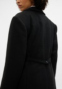 Černý kabát z vlněné směsi s strukturovaným designem, s klopovým límcem, detaily s knoflíkem na zadní straně a jemnou texturou. K tailored střih se zvýrazněnými ramenními vycpávkami.