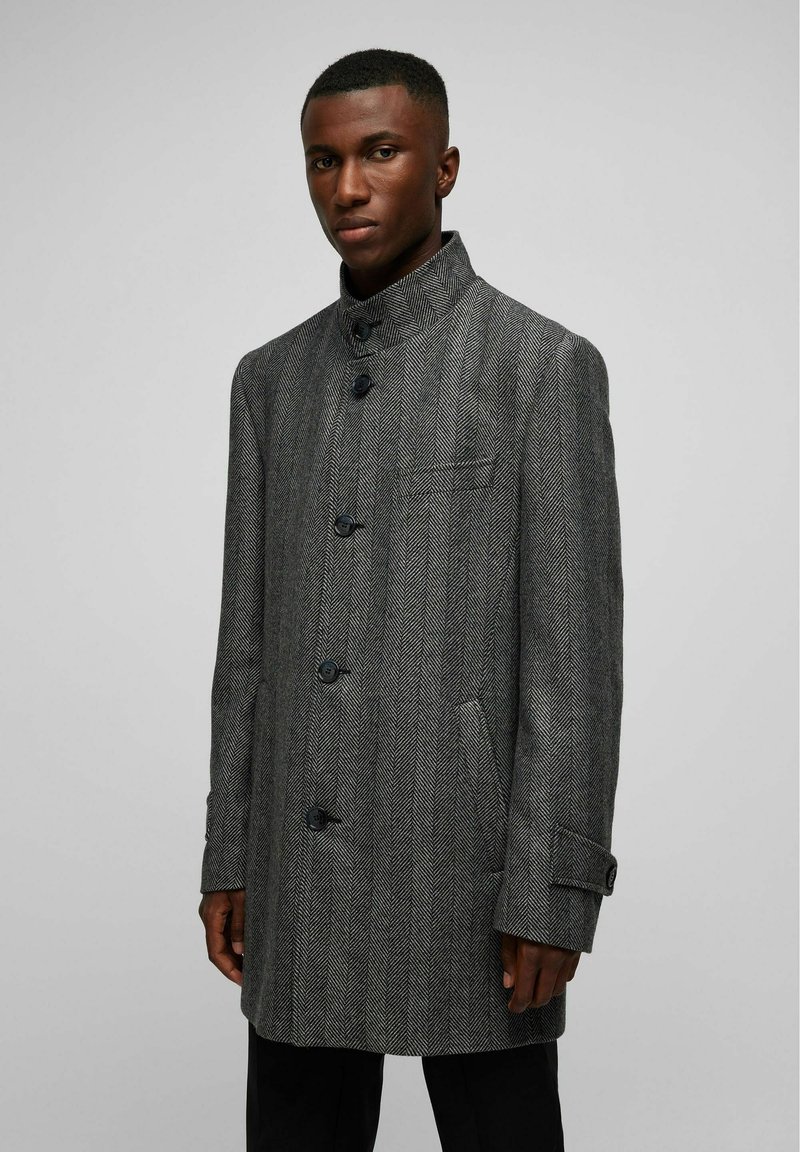 Manteau en chevrons gris avec un col haut, six boutons noirs, deux poches latérales et une petite poche poitrine, fabriqué à partir d'un tissu texturé.