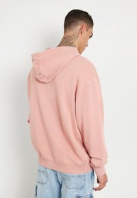 Rožinė medvilninė džemperis su raištelių gobtuvu, laisvas kirpimas ir banguoti rankogaliai. Pasižymi vientisa spalva ir minkšta tekstūra. Rodymas iš nugaros.