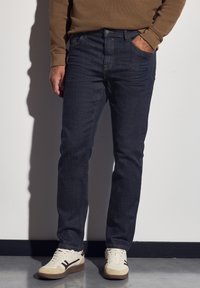 Hombre con jeans azul oscuro, camisa de manga larga marrón y zapatillas blancas de pie contra una pared blanca con una mano en el bolsillo.