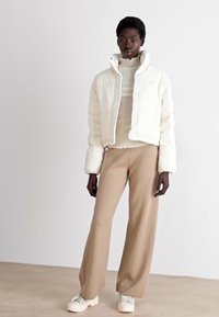 Doudoune off-white, au design court avec une finition brillante. Associée à un pantalon large beige et des baskets blanches. Tissu texturé.