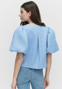 Ljusblå puffärmad topp i textil med strukturerad yta, med fyrkantig halsringning och slits i ryggen. Matchas med figurnära blå jeans.