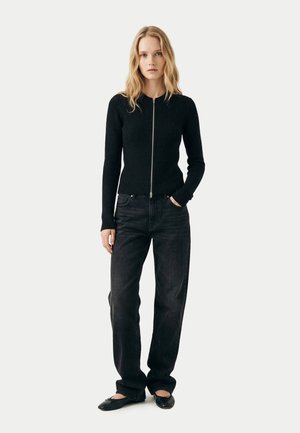 Blonde Frau trägt einen schwarzen Reißverschluss-Pullover, schwarze Jeans und schwarze Ballerinas, steht mit einer Hand in der Tasche.
