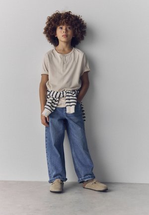 Child with curly hair stands against a white wall wearing a beige shirt, striped sweater tied at the waist, blue jeans, and beige slip-on shoes.