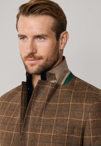 Brauner Tweed-Mantel mit Karomuster, der einen hohen Kragen mit grünen Akzenten und einem dunklen Innenfutter aufweist, das eine strukturierte Oberfläche betont.