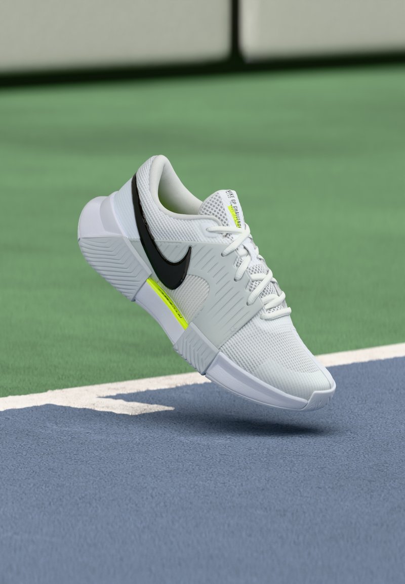 Λευκό αθλητικό παπούτσι με μαύρες λεπτομέρειες, πλεκτό υλικό, υφή στην σόλα και φωτεινή κίτρινη λεπτομέρεια. Τοποθετημένο πάνω από μπλε επιφάνεια γηπέδου.